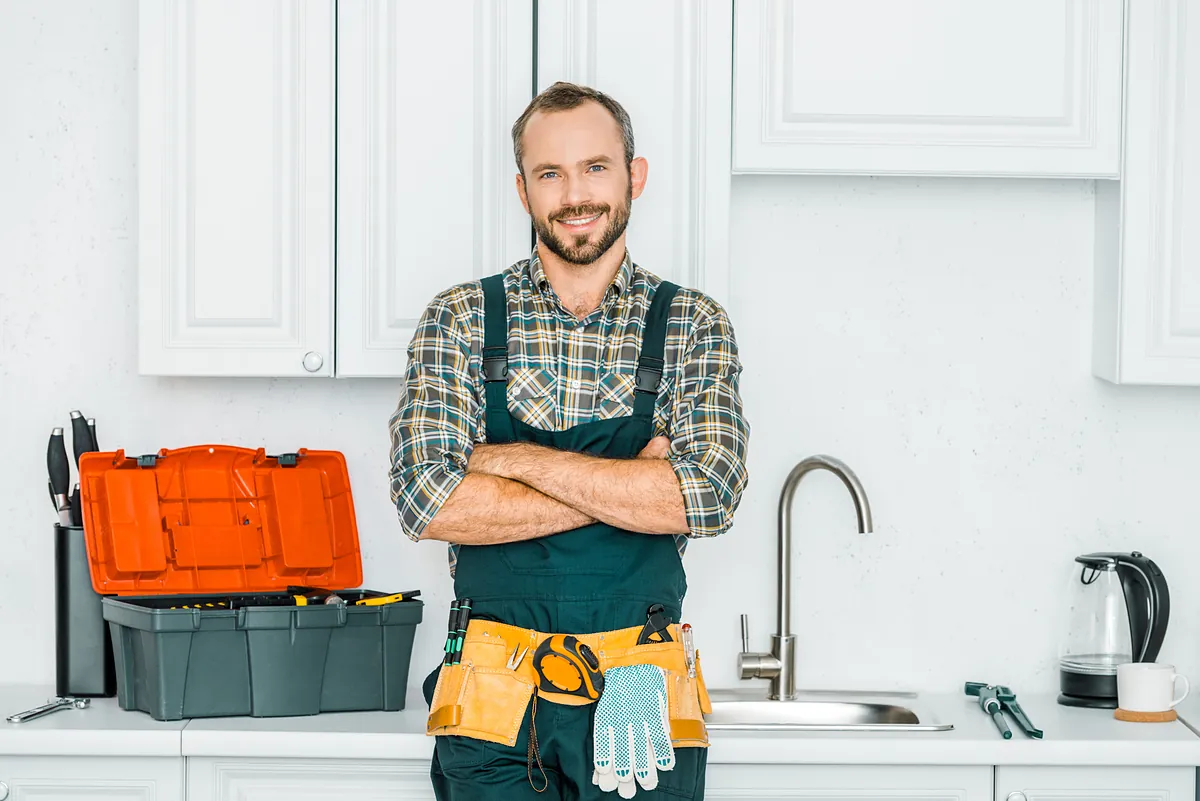 Installateur für Rohrreinigung mit Werkzeugkoffer in der Küche Freundlicher Installateur mit Werkzeugkoffer und Werkzeuggürtel in einer Küche